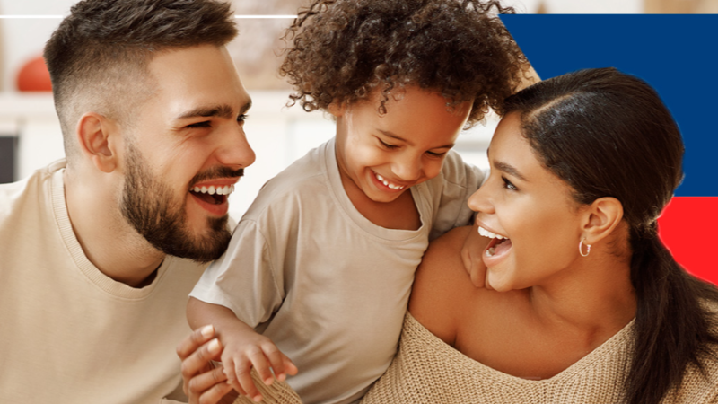 Homem, mulher e criança sorriem juntos em ambiente interno. A criança está ao centro, interagindo com os adultos, enquanto os três demonstram um momento de convivência e alegria.
