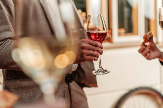 Pessoa segura uma taça de vinho rosé durante um momento de confraternização. Ao fundo, outras taças e participantes aparecem desfocados, criando uma atmosfera associada à degustação e à experiência enogastronômica.