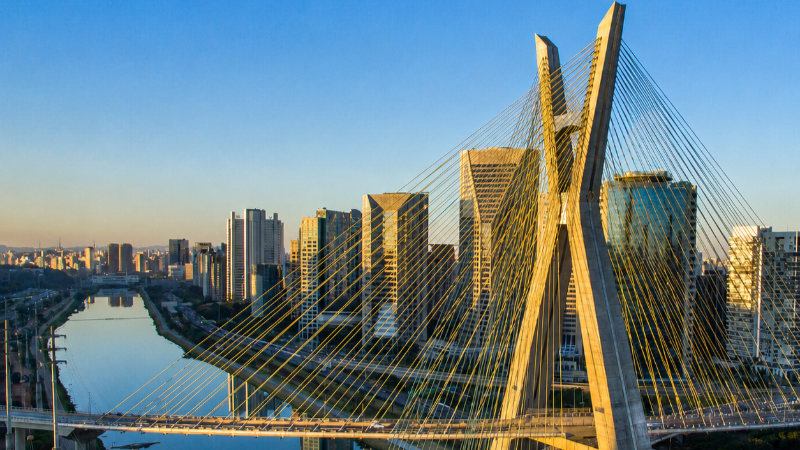 Imagem aérea da Ponte Estaiada sobre o Rio Pinheiros durante o pôr do sol, com prédios corporativos ao fundo e reflexos na água.