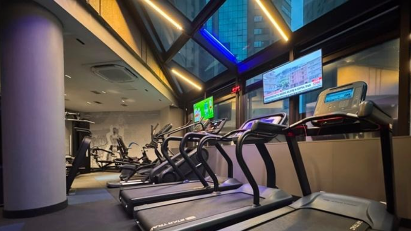 Academia do Wyndham São Paulo Ibirapuera com esteiras alinhadas em primeiro plano, além de bicicletas ergométricas e outros equipamentos de musculação ao fundo. O ambiente possui amplas janelas de vidro com vista para edifícios da cidade, iluminação em LED e televisores instalados acima dos aparelhos.