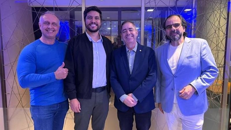 Quatro homens posam lado a lado em um ambiente interno do Wyndham São Paulo Ibirapuera. Três deles vestem blazer ou paletó, enquanto o primeiro, à esquerda, usa camiseta azul de manga longa e faz um gesto de positivo com a mão. Todos sorriem para a câmera. Ao fundo, há portas de vidro, iluminação em tons azulados e elementos decorativos que remetem ao lobby do hotel.